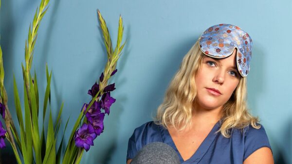 Person wearing a patterned brocade sleep mask beside tall purple flowers against a blue wall