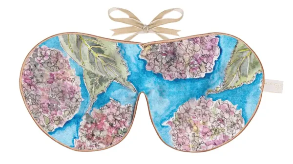 Silk sleep mask featuring a blue background with illustrated hydrangea blooms in pink, lavender, and cream tones, framed with warm piping and finished with a beige ribbon tie.