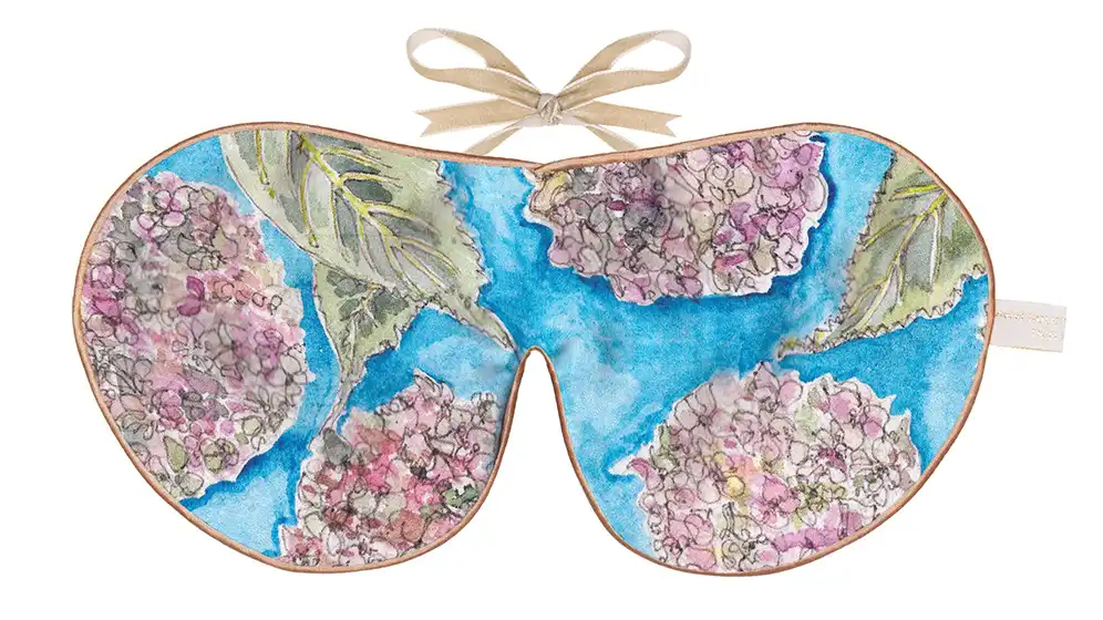 Silk sleep mask featuring a blue background with illustrated hydrangea blooms in pink, lavender, and cream tones, framed with warm piping and finished with a beige ribbon tie.