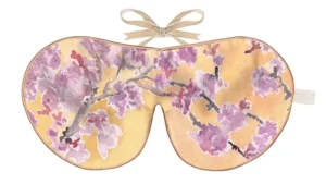 The image shows a silk sleep mask decorated with a botanical illustration inspired by hydrangea flowers. The front surface displays clusters of rounded hydrangea blossoms positioned across the mask in soft shades of pink, lavender, lilac, and pale cream. Each cluster is composed of many small petal shapes, rendered with layered watercolour-style shading and fine ink outlines that give a hand‑drawn, detailed effect. Between the hydrangea blooms are large, elongated leaves in varied shades of green, with textured vein lines that add depth. The background is a bright, clear blue wash, creating a vivid contrast against the softer floral colours and giving the design a fresh, summery quality. The mask is finished with warm-toned piping along the edge, outlining the curved silhouette. At the top, a beige ribbon is tied into a neat bow, indicating a ribbon‑tie fastening. A small fabric tag is attached at the right edge. The overall look is vibrant, botanical, and painterly, with an emphasis on floral detail and colour harmony.