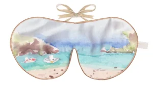 Silk sleep mask with a coastal scene showing turquoise water, sandy shoreline, and distant cliffs in soft watercolour tones.