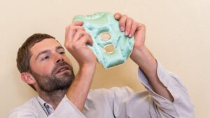 Head and shoulder shot of a man stood up holding a Jade Holistic Silk Mulberry Silk Face Pillow weighted full face pillow up in the air prior to putting it on