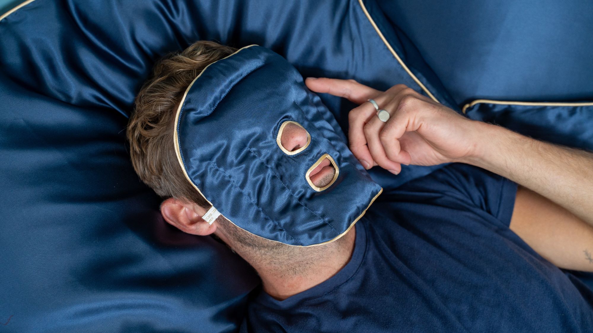 Head and shoulder shot of a man laid on a navy pillowcase holding a Holistic Silk Navy Mulberry Silk weighted full face pillow on his face.
