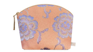 A flat image of the Holistic Silk Lavender Make Up Bag in a Peach and Lilac colour abstract floral brocade on a white background.. The make up bag is stood up and is wider at the top than the bottom, it has a curved zipped top which has a honey colour piping outlining it and the zip pull hanging to the top left of the bag.