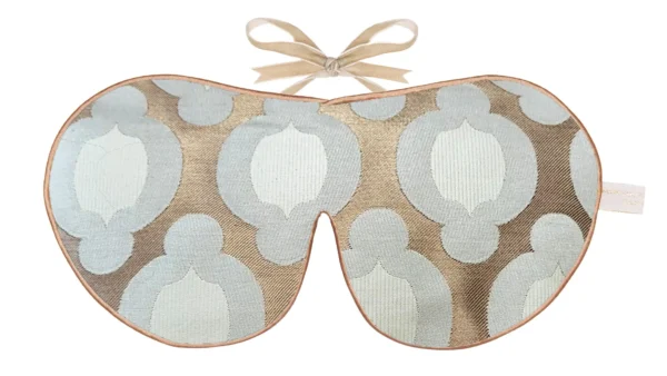 A silk sleep mask with a geometric woven pattern in soft blue and taupe tones, finished with gold edging and beige ribbon ties.