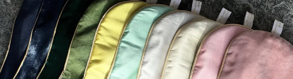 A row of silk sleep masks arranged in a gradient of colours from deep navy to pastel pink, each with gold trim and fabric loops.”