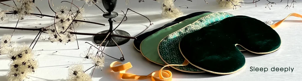 A group of green and patterned silk sleep masks arranged beside delicate dried botanical stems, with the text ‘Sleep deeply.