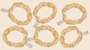 Six cream satin silk scrunchies arranged on a warm beige background, each with a white fabric tag.
