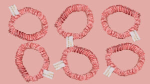Six rose-pink satin silk scrunchies displayed on a pink background, each with a small white tag.