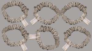 Six silver satin silk scrunchies arranged on a light grey background, each with a white tag attached.