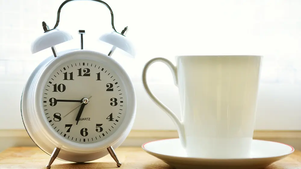 White analog alarm clock next to a white cup and saucer on a wooden surface, with soft natural light coming from behind.