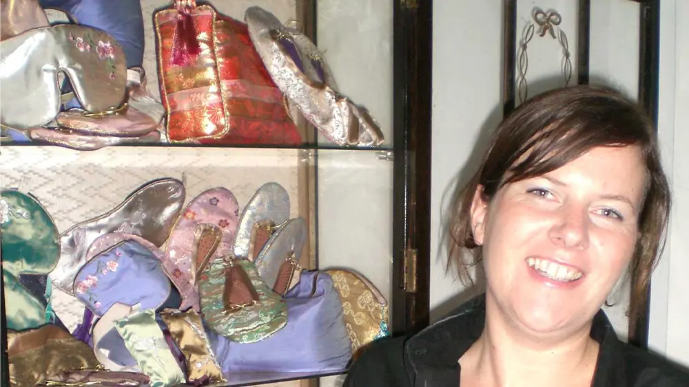 A person in a dark collared top stands next to a glass display cabinet filled with colorful silk accessories, including slippers, pouches, and eye masks.