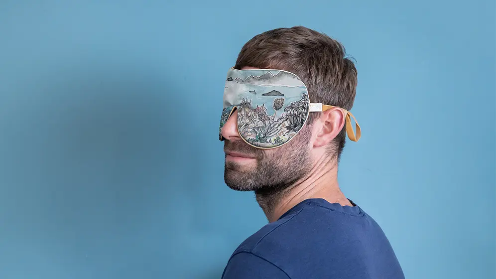 Model wearing Coastal Succulents exclusive silk sleep mask with honey‑coloured trim against a soft blue background