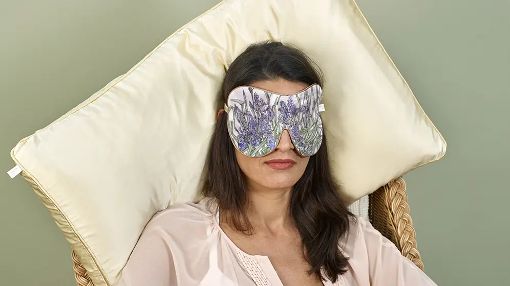 Person reclining wearing Iris exclusive silk sleep mask with honey trim on a cream cushion