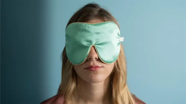 Jade silk sleep mask worn over the eyes, finished with honey‑coloured piping, shown from the front against a soft blue background.