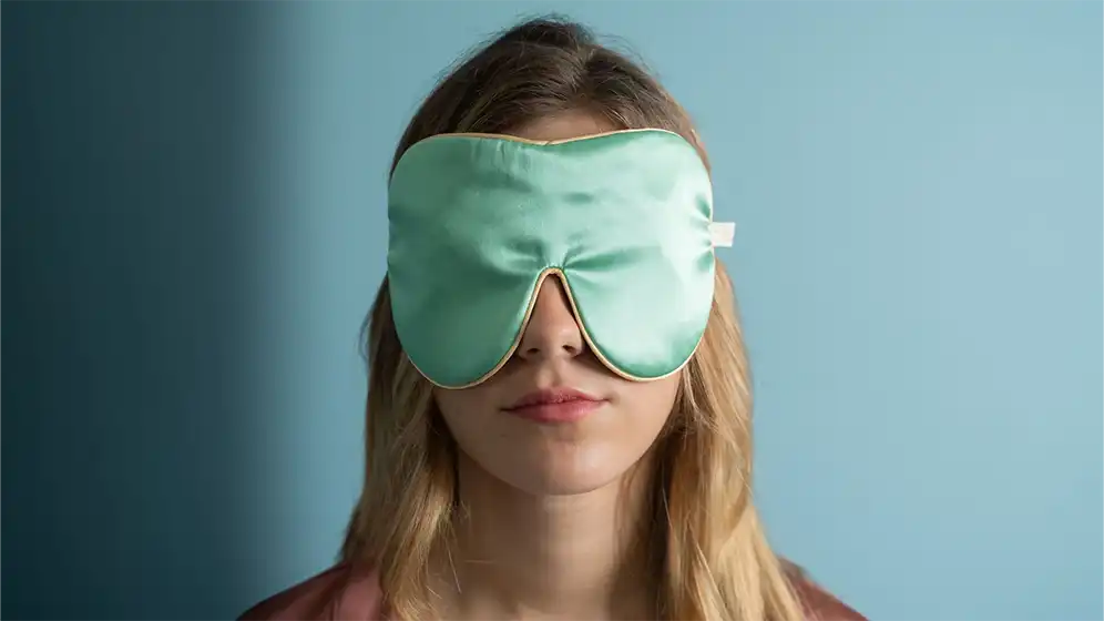 Jade silk sleep mask worn over the eyes, finished with honey‑coloured piping, shown from the front against a soft blue background.