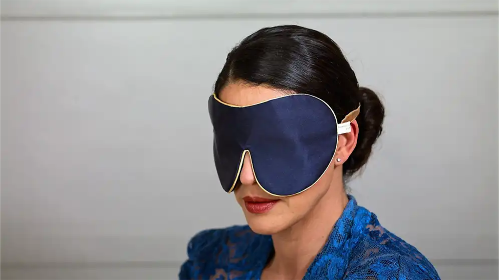 Navy pure silk one strap sleep mask with honey coloured piping worn on a model, shown in side view against a neutral background.