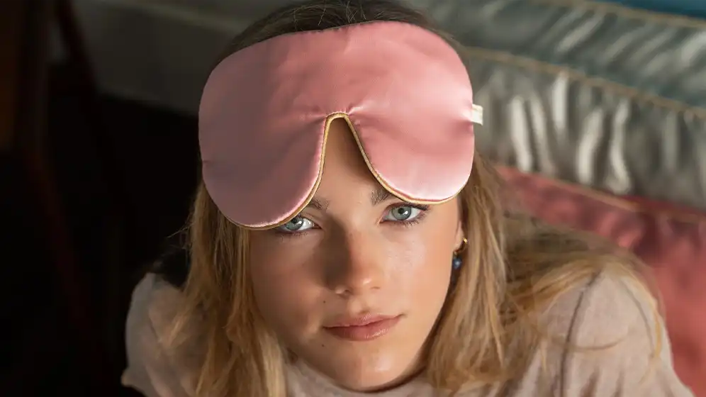 Rose silk sleep mask worn above the eyes, finished with honey‑coloured piping, shown in a close‑up lifestyle setting.