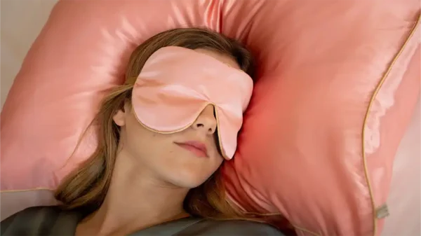 Rose silk sleep mask worn while resting on a matching rose silk pillow with honey‑coloured piping, shown in a close‑up lifestyle setting.