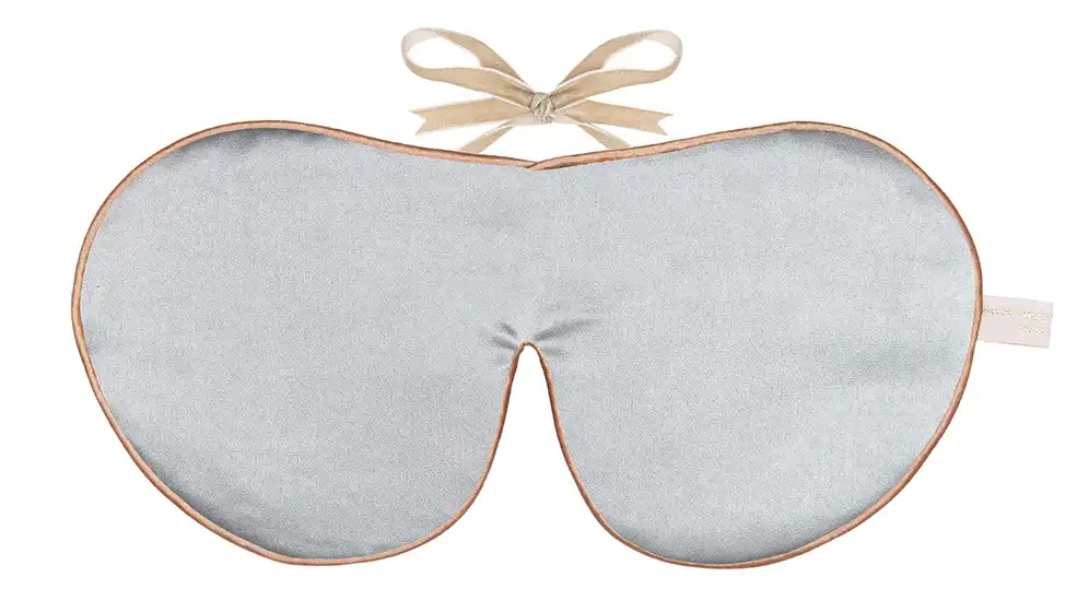 A silver silk sleep mask is displayed flat against a white background. The mask is made from silk and has a smooth surface with a soft reflective sheen. The shape is wide and contoured to fit over the eyes, with a curved centre section for the bridge of the nose.