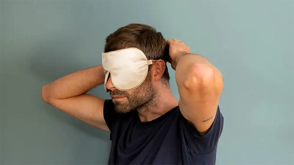 Silver silk sleep mask being secured at the back of the head with a silk band, shown from a rear three‑quarter view against a soft green background.